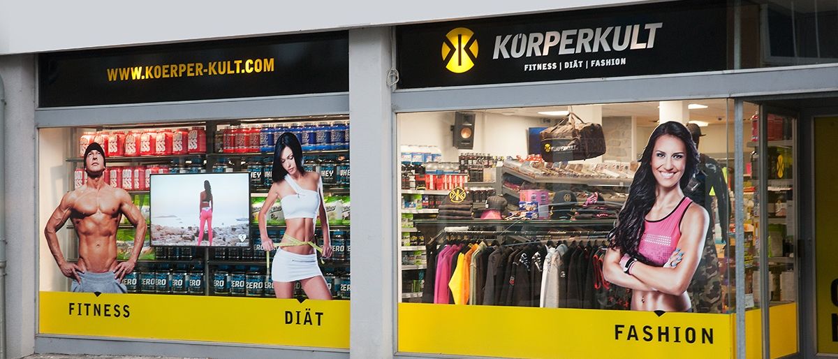 Fassade eines Fitness- und Modegeschäfts. Zeigt Fitnessgetränke, eine Frau mit Maßband und eine lächelnde Frau.