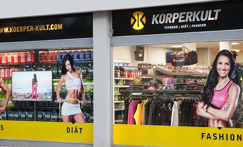 Fassade eines Fitness- und Modegeschäfts. Zeigt Fitnessgetränke, eine Frau mit Maßband und eine lächelnde Frau.