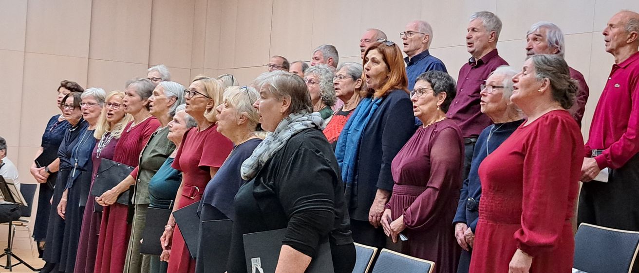 Ein Chor älterer Erwachsener, einige mit Brille, in formeller Kleidung, singen in einem Raum mit beigen Wänden und Stühlen.