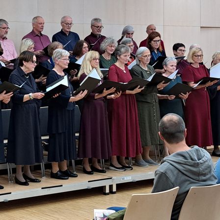 Ein Chor von Menschen in formeller Kleidung singt auf der Bühne mit Musikständen in einem Raum mit Holzböden und einem gelben Banner mit Text auf der linken Seite.