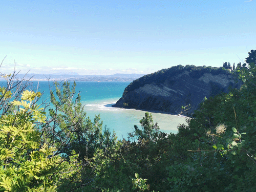 Ein Blick von einem Hügel zeigt ein ruhiges Meer mit einer Klippe auf der rechten Seite unter einem klaren blauen Himmel.