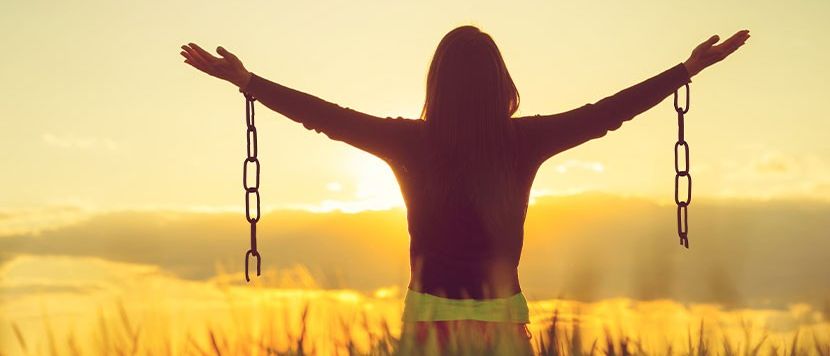 Eine Frau mit ausgestreckten Armen in einem Feld mit Ketten an den Handgelenken, die in Richtung Sonnenuntergang schaut.