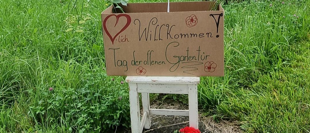 Eine Pappschachtel mit einem Herz und dem Text 'Ich Will Kommen' steht auf einem weißen Hocker in einem Garten.
