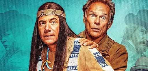 Zwei Männer in Kostümen, einer als Indianer und der andere als Cowboy gekleidet, stehen eng beieinander vor einem blauen Hintergrund.