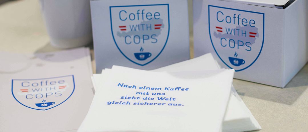 Eine weiße Tasse und zwei Schachteln mit der Aufschrift 'Coffee with Cops' mit blauem Text auf weißem Hintergrund. Ein Seidenpapier mit einem Zitat über Kaffee und Sicherheit.