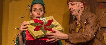 Eine Frau in einer Weihnachtsmütze öffnet ein verpacktes Geschenk, während ein Mann in einem Kegelhut zuschaut, beide vor einer gelben Wand mit Weihnachtslichtern.