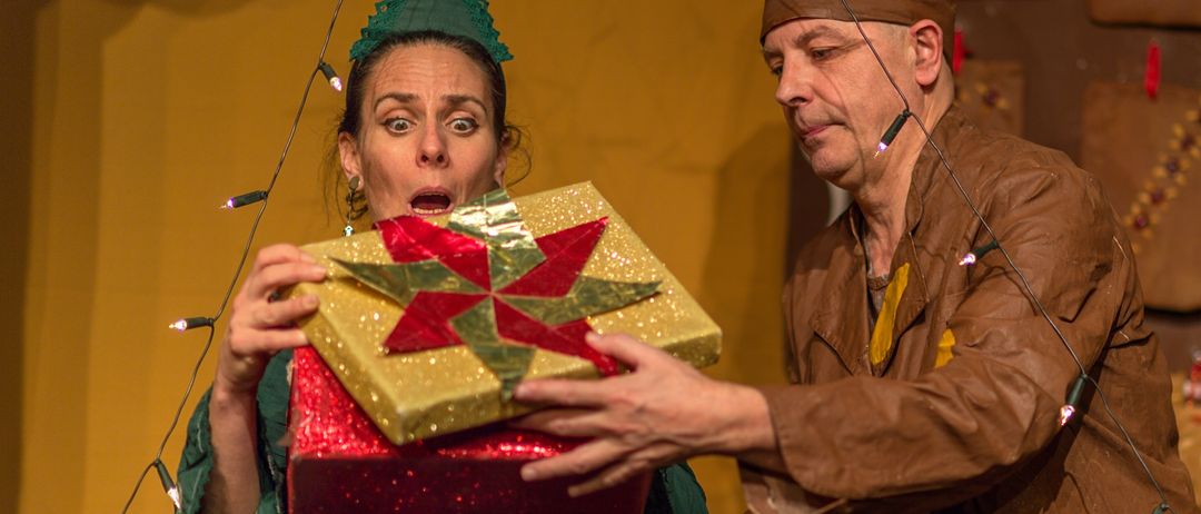 Eine Frau in einer Weihnachtsmütze öffnet ein verpacktes Geschenk, während ein Mann in einem Kegelhut zuschaut, beide vor einer gelben Wand mit Weihnachtslichtern.