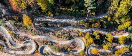 Eine Luftaufnahme einer kurvenreichen Erdstraße durch ein bewaldetes Gebiet, umgeben von einer Mischung aus grünem und gelbem Laub, mit dem Schatten einer Person auf dem Boden sichtbar.