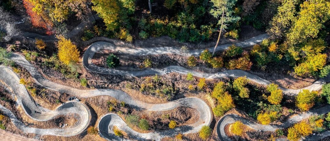 Eine Luftaufnahme einer kurvenreichen Erdstraße durch ein bewaldetes Gebiet, umgeben von einer Mischung aus grünem und gelbem Laub, mit dem Schatten einer Person auf dem Boden sichtbar.