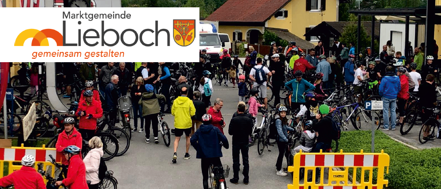 Eine große Menschenmenge in verschiedenen Outfits und Helmen versammelt sich um Fahrräder. Sie befinden sich in einem öffentlichen Raum mit Häusern und einem Uhrturm im Hintergrund. Ein Schild im Bild lautet 'Marktgemeinde Lieboch'.