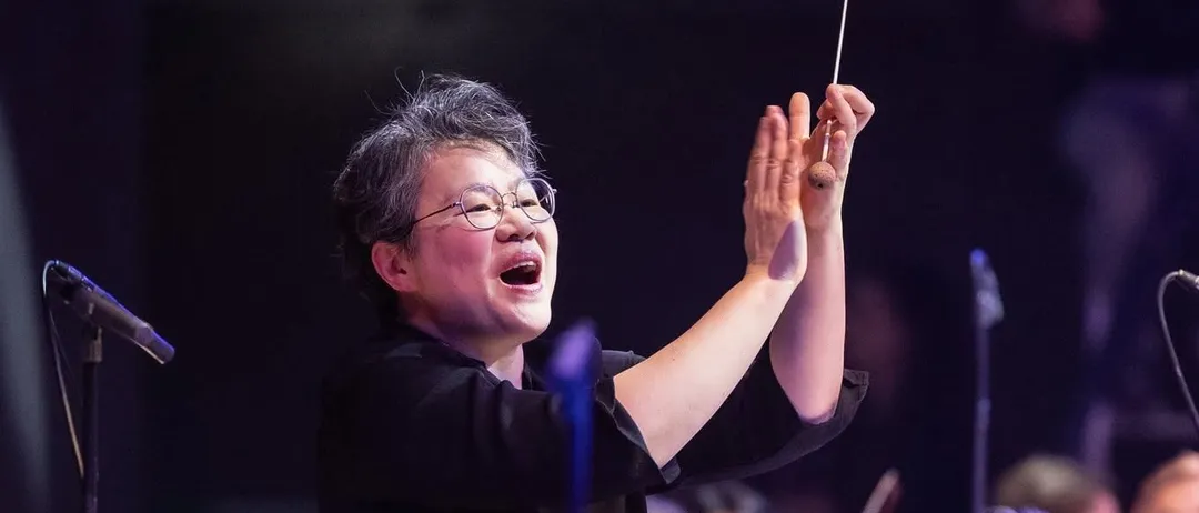 Ein Dirigent leitet ein Orchester während einer Aufführung mit Publikum im Hintergrund, ein Violinist hält sein Instrument und die Sicht auf das Publikum ist verschwommen.