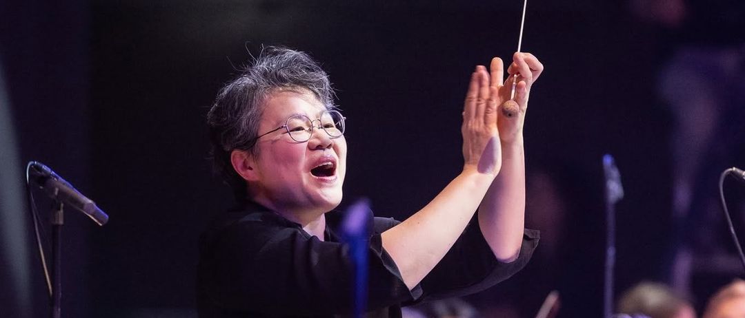 Ein Dirigent leitet ein Orchester während einer Aufführung mit Publikum im Hintergrund, ein Violinist hält sein Instrument und die Sicht auf das Publikum ist verschwommen.