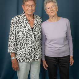 Zwei ältere Frauen stehen nebeneinander und lächeln. Die Frau auf der linken Seite trägt einen Leopardenmuster-Blazer und die Frau auf der rechten Seite ein lilafarbenes Oberteil und schwarze Hose. Beide tragen eine Brille.