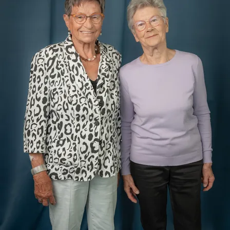 Zwei ältere Frauen stehen nebeneinander und lächeln. Die Frau auf der linken Seite trägt einen Leopardenmuster-Blazer und die Frau auf der rechten Seite ein lilafarbenes Oberteil und schwarze Hose. Beide tragen eine Brille.