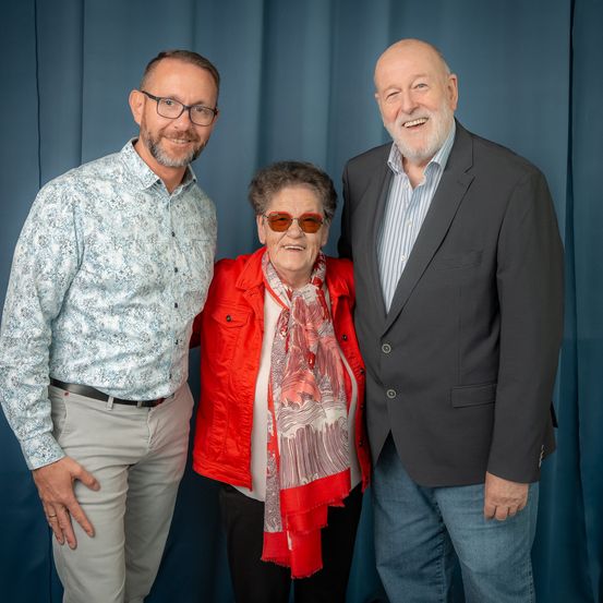 Drei Personen stehen vor einem blauen Hintergrund. Der Mann links trägt eine Brille und ein gemustertes Hemd, die Frau in der Mitte hat eine rote Jacke und einen Schal, und der Mann rechts trägt einen grauen Anzug.
