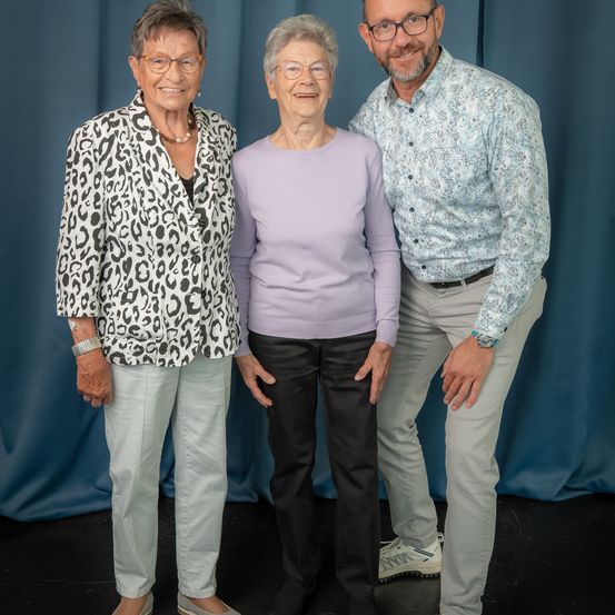 Drei Personen, zwei Frauen und ein Mann, stehen eng zusammen in einem Studio mit blauem Hintergrund. Die Frauen tragen Brillen, und eine trägt eine Leoparden-Jacke. Der Mann trägt ein gemustertes Hemd.