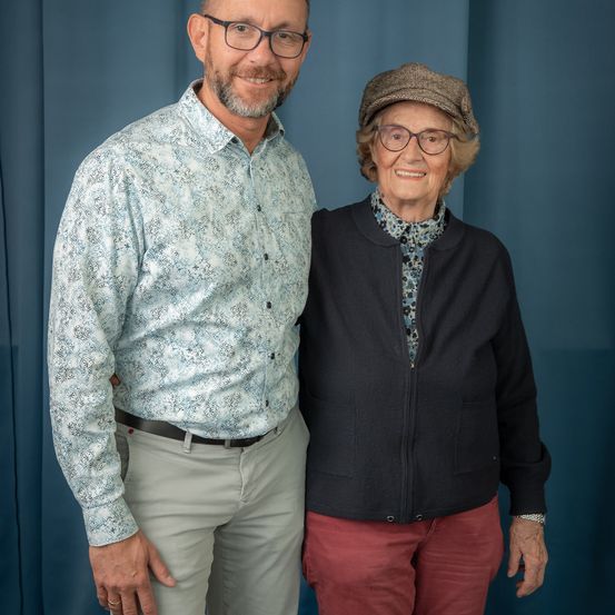 Ein Mann und eine ältere Frau stehen nebeneinander und lächeln für ein Foto. Der Mann trägt eine Brille und ein Blumenhemd, während die Frau eine Mütze und eine schwarze Strickjacke trägt.