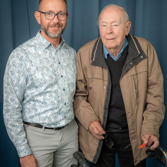 Zwei Männer stehen zusammen vor einem blauen Hintergrund. Der jüngere Mann trägt ein gemustertes Hemd und eine Brille und lächelt. Der ältere Mann hat graues Haar, trägt eine braune Jacke und hält eine Brille.