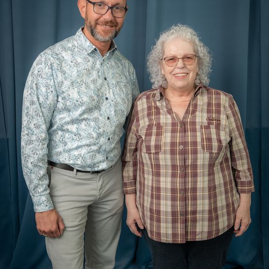 Ein Mann und eine Frau stehen lächelnd nebeneinander vor einem blauen Vorhang. Der Mann trägt ein gemustertes Hemd und eine Hose, während die Frau ein kariertes Hemd trägt. Beide tragen eine Brille.