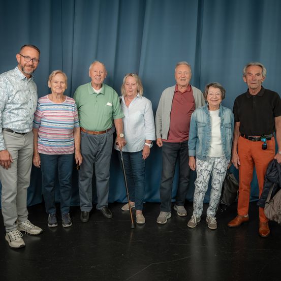 Eine Gruppe von sieben älteren Personen, darunter Männer und Frauen, posiert für ein Foto vor einem blauen Vorhang. Sie alle lächeln.