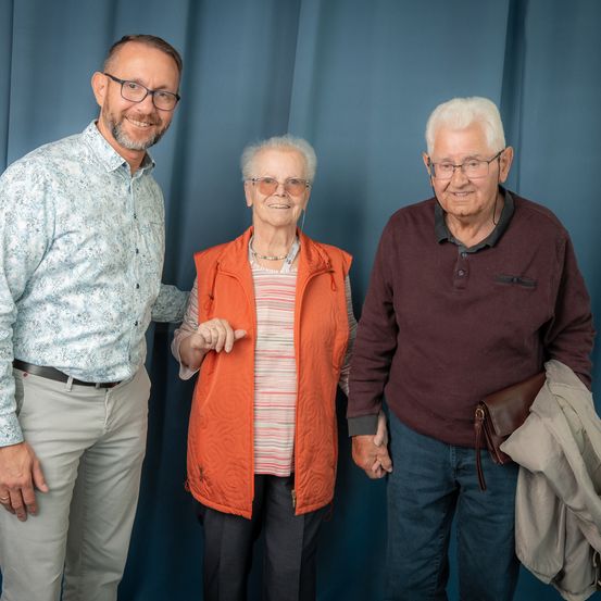 Drei Menschen, zwei Männer und eine Frau, stehen lächelnd vor einem blauen Vorhang. Die Frau trägt eine orange Weste und der Mann auf der rechten Seite hält eine Jacke.