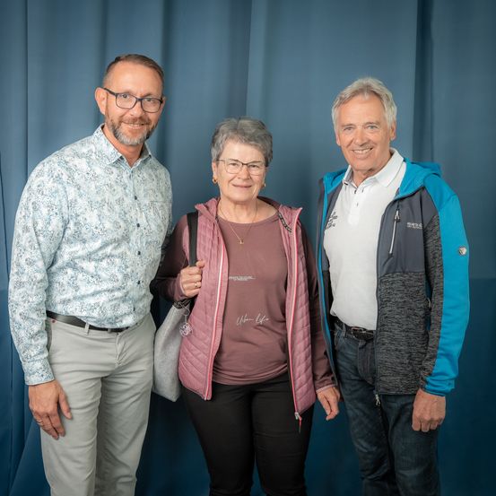 Drei Menschen stehen vor einem blauen Vorhang. Der Mann links trägt ein gemustertes Hemd und eine Brille. Die Frau in der Mitte hat eine Halskette und hält eine Tasche. Der Mann rechts trägt eine Jacke mit Kapuze und ein weißes Hemd.