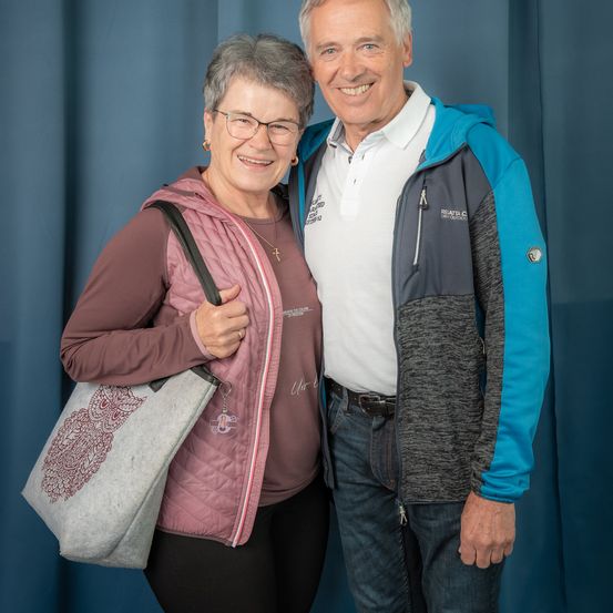 Ein älteres Paar steht eng beieinander und lächelt hell vor einem blauen Hintergrund. Die Frau trägt eine Brille und eine rosa Weste, während der Mann eine blaue Jacke trägt.