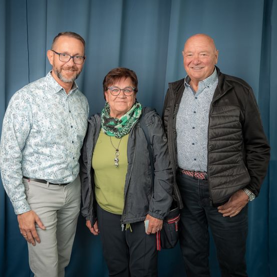Drei Personen stehen eng beieinander vor einem blauen Vorhang. Der Mann auf der linken Seite trägt eine Brille und ein Hemd mit Blumenmuster. Die Frau in der Mitte trägt eine Brille und einen Schal. Der Mann auf der rechten Seite trägt eine schwarze Jacke.