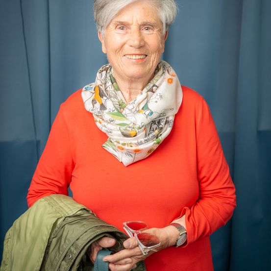 Eine ältere Frau mit weißen Haaren lächelt, trägt ein rotes Langarmshirt und einen Blumenschal. Sie hält eine grüne Jacke und eine Sonnenbrille. Hinter ihr ist ein blauer Vorhang.