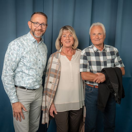 Drei Menschen stehen nebeneinander vor einem blauen Vorhang. Der Mann links trägt eine Brille und ein gemustertes Hemd, die Frau in der Mitte trägt eine Halskette und eine karierte Jacke und der Mann rechts hält einen Mantel und trägt ein kariertes Hemd.