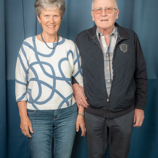 Ein älteres Paar steht lächelnd eng beieinander vor einem blauen Vorhang. Die Frau trägt ein weiß-blau gemustertes Oberteil und Jeans. Der Mann trägt eine schwarze Jacke über einem karierten Hemd.