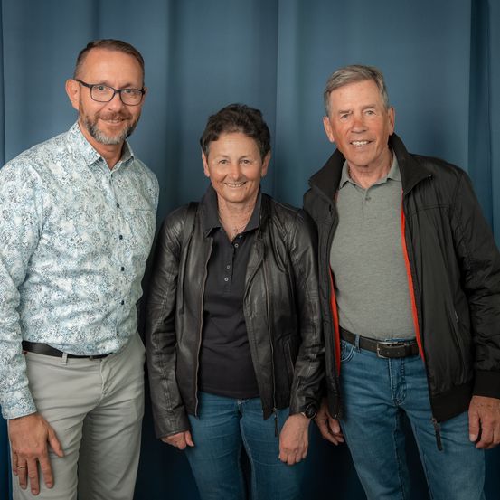 Drei Menschen stehen eng beieinander vor einem blauen Vorhang und lächeln für ein Foto. Der Mann links trägt eine Brille und ein gemustertes Hemd, die Frau in der Mitte eine schwarze Lederjacke und der Mann rechts ein graues Hemd unter einer schwarzen Jacke.