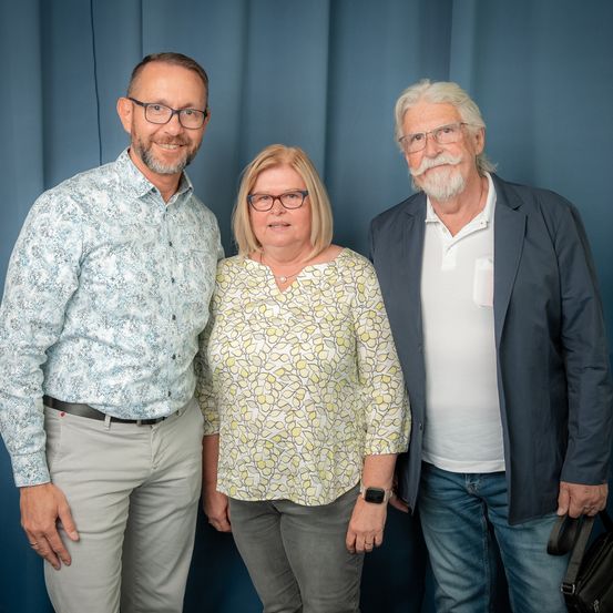 Drei Personen stehen gemeinsam vor einem blauen Hintergrund. Ein Mann links trägt ein gemustertes Hemd und eine Brille. In der Mitte lächelt eine Frau mit Brille und einer Halskette. Der Mann rechts hat einen weißen Bart, trägt eine Anzugsjacke und hält eine schwarze Tasche.