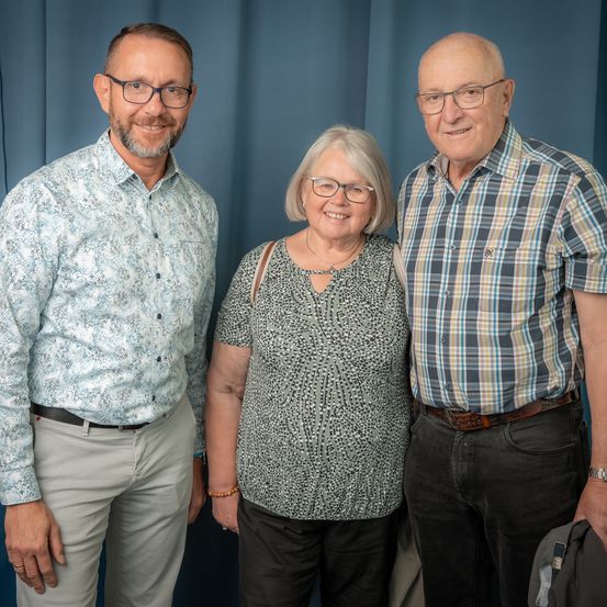 Drei Menschen stehen zusammen vor einem blauen Vorhang. Der Mann auf der linken Seite trägt eine Brille und einen Gürtel, die Frau in der Mitte hat eine Halskette und ein Armband, und der Mann auf der rechten Seite trägt ein kariertes Hemd.