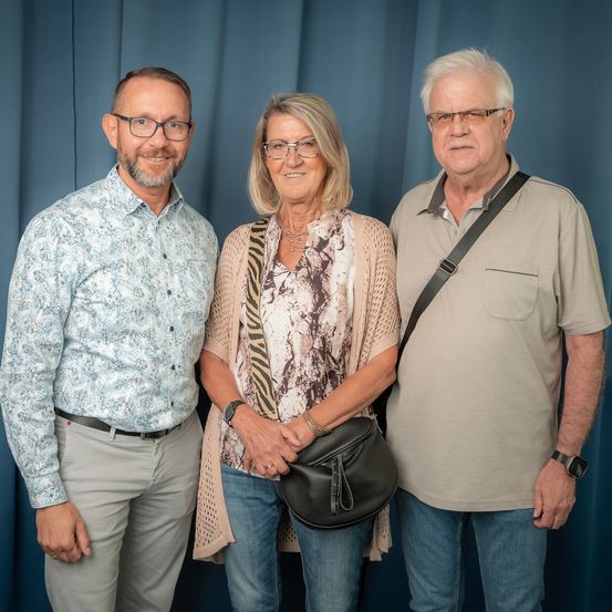 Drei Personen stehen nebeneinander vor einem blauen Vorhang. Der Mann auf der linken Seite trägt ein Blumenhemd und eine Brille. Die Frau in der Mitte hat eine Handtasche und trägt eine Halskette. Der Mann auf der rechten Seite trägt ein beiges Poloshirt und eine schwarze Umhängetasche.