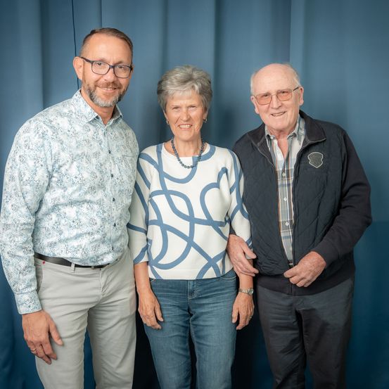 Drei Menschen stehen eng beieinander und lächeln. Eine Frau in der Mitte trägt eine Halskette. Der Mann rechts trägt eine schwarze Jacke.