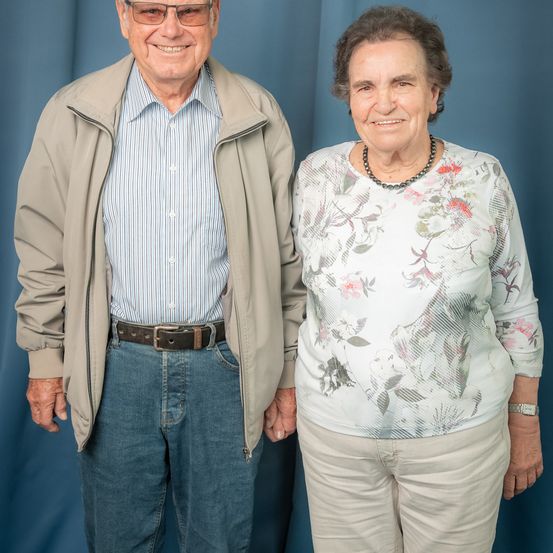 Ein älterer Mann und eine ältere Frau stehen lächelnd vor einem blauen Hintergrund. Der Mann trägt eine Brille, eine Jacke und ein gestreiftes Hemd. Die Frau trägt eine Bluse mit Blumenmuster und eine Halskette.