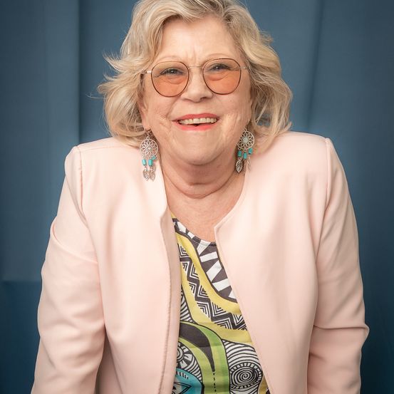 Eine ältere Frau mit blonden Haaren lächelt. Sie trägt eine Brille, einen rosa Blazer und türkisfarbene Ohrringe. Sie steht vor einem blauen Hintergrund.