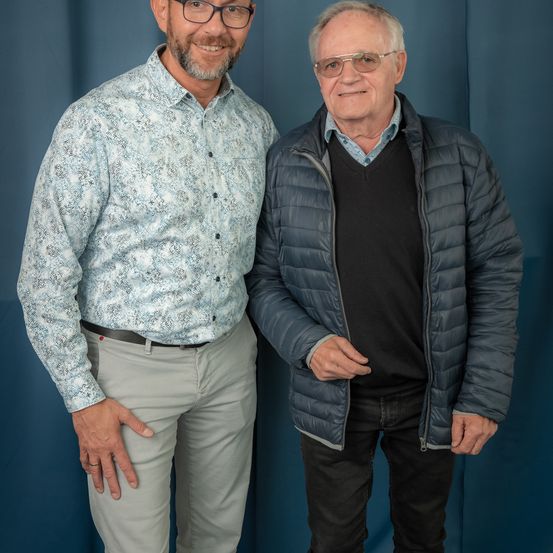 Zwei Männer stehen eng beieinander und lächeln für ein Foto. Der Mann auf der linken Seite trägt eine Brille und ein gemustertes Hemd. Der Mann auf der rechten Seite trägt ein schwarzes Shirt unter einer gefütterten Jacke.