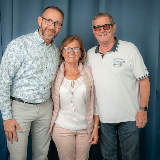 Drei Personen stehen gemeinsam für ein Foto. Der Mann links trägt ein gemustertes Hemd und eine Brille. Die Frau in der Mitte trägt eine Halskette und eine Brille. Der Mann rechts trägt ein weißes Poloshirt und eine Sonnenbrille. Sie alle lächeln.