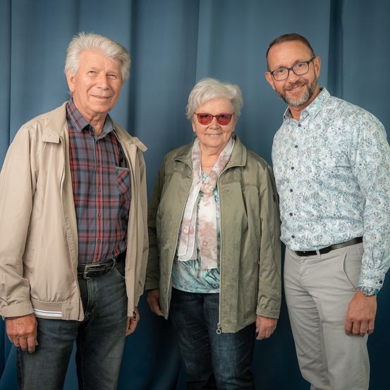 Drei Personen stehen dicht beieinander vor einem blauen Vorhang. Der Mann auf der linken Seite trägt eine helle Jacke und ein kariertes Hemd. Die Frau in der Mitte hat weißes Haar und trägt eine Sonnenbrille. Der Mann auf der rechten Seite trägt eine Brille und ein gemustertes Hemd.