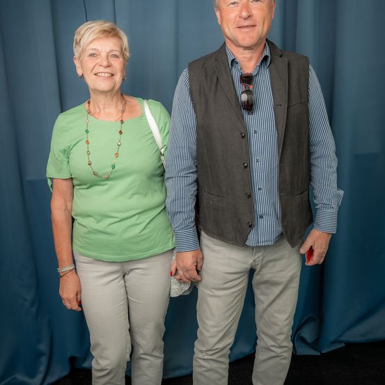 Eine ältere Frau und ein Mann stehen nah beieinander und lächeln. Die Frau trägt ein grünes Shirt und eine Halskette, der Mann ein blaues Hemd mit einer Weste. Dahinter ist ein blauer Vorhang.