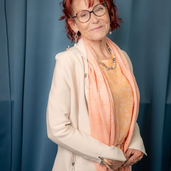 Eine ältere Frau mit roten Haaren und Brille lächelt, während sie vor einem blauen Vorhang steht. Sie trägt eine helle Jacke, ein Blusenhemd mit Blumenmuster und eine Perlenkette.