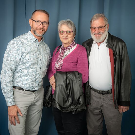 Drei Personen stehen nebeneinander vor einem blauen Hintergrund. Zwei Männer tragen Brillen, einer eine Lederjacke. Die Frau trägt einen Schal und hält eine Ledertasche.