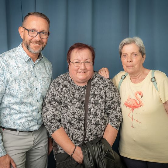 Drei Personen, zwei Frauen und ein Mann, stehen gemeinsam vor einem blauen Hintergrund. Der Mann trägt eine Brille und ein gemustertes Hemd. Die Frau in der Mitte trägt eine Brille und ein gemustertes Oberteil. Die Frau auf der rechten Seite trägt ein gelbes T-Shirt mit einem Flamingo-Design.