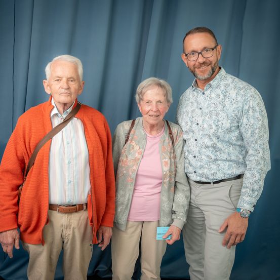 Drei Personen stehen eng zusammen und lächeln für ein Foto. Der Mann links trägt einen orangefarbenen Pullover, die Frau in der Mitte hat eine Jacke mit Blumenmuster und der Mann rechts trägt ein Hemd mit Blumenmuster. Sie tragen alle helle Hosen. Hinter ihnen ist ein blauer Vorhang.