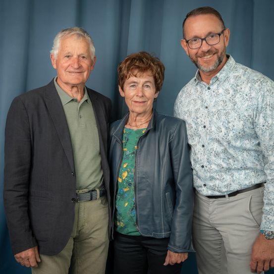 Drei Personen, zwei Männer und eine Frau, stehen eng nebeneinander vor einem blauen Hintergrund und lächeln für ein Foto.