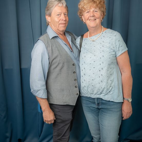 Ein älteres Paar steht lächelnd eng nebeneinander, mit einem blauen Vorhang als Hintergrund. Der Mann trägt eine graue Weste über einem blauen Hemd, und die Frau trägt ein hellblaues Oberteil mit Muster und Jeans.