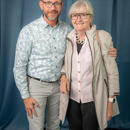 Ein Mann und eine Frau stehen eng beieinander und lächeln. Der Mann trägt ein Blumenhemd und Hose, während die Frau eine beige Jacke und eine Halskette trägt. Sie stehen vor einem blauen Hintergrund.
