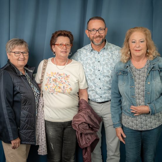 Vier Personen stehen vor einem blauen Vorhang. Zwei Frauen tragen Brillen, eine hält eine Jacke. Der Mann trägt eine Brille und ein gemustertes Hemd. Die vierte Frau hat blonde Haare und trägt eine Jeansjacke.